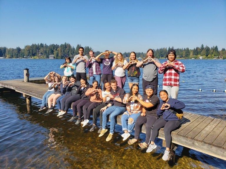 Students at the New Student Retreat sit and stand on a dock stretching out into the lake, making hearts with their hands on a sunny day