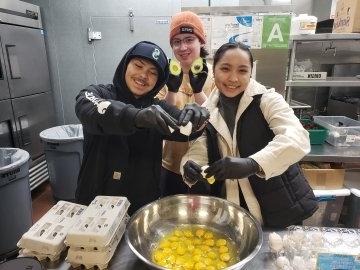 Students crack eggs at the community kitchen into a giant metal bowl full of eggs; one student holds up an avocado cut in half